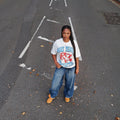 Person standing on a road with white markings, wearing a white t-shirt with red and blue designs.