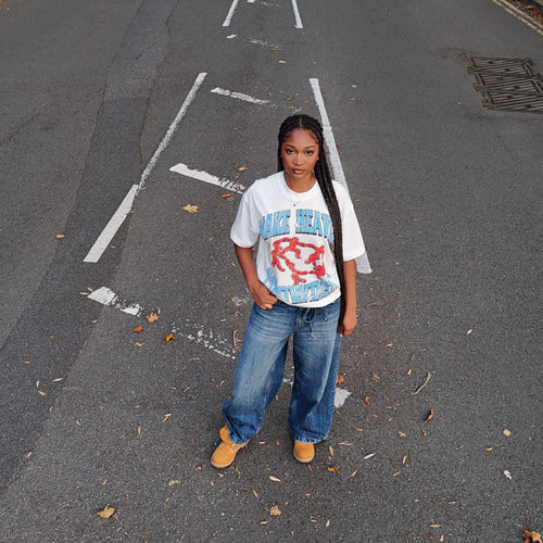 Person standing on a road with white markings, wearing a white t-shirt with red and blue designs.